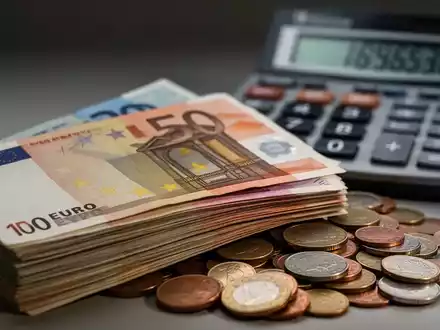 Ein Stapel Euro-Banknoten, teilweise verdeckt von Kleingeld, mit einem Taschenrechner im unscharfen Hintergrund, der steigende Zahlen anzeigt. - Foto: über boerse-global.de