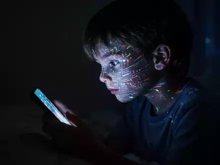 Ein Kind, dessen Gesicht vom leuchtenden Bildschirm eines Smartphones beleuchtet wird, sitzt isoliert in einem dunklen Raum. - Foto: über boerse-global.de