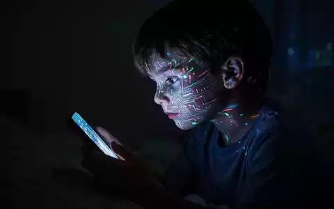 Ein Kind, dessen Gesicht vom leuchtenden Bildschirm eines Smartphones beleuchtet wird, sitzt isoliert in einem dunklen Raum. - Foto: über boerse-global.de