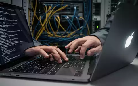 Nahaufnahme von Händen, die auf einer MacBook-Tastatur in einer IT-Umgebung tippen, mit Server-Racks im unscharfen Hintergrund. Auf dem Bildschirm ist Code zu sehen. - Foto: über boerse-global.de