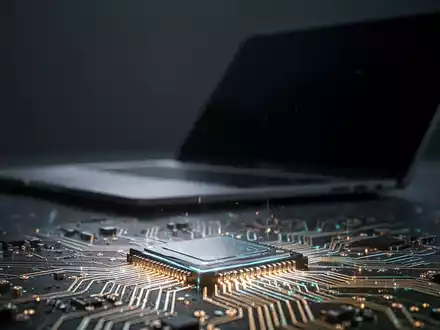 Nahaufnahme eines leuchtenden Mikrochips mit feinen Schaltkreisen, im unscharfen Hintergrund dezent ein Laptop angedeutet. - Foto: über boerse-global.de