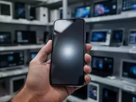 Eine Hand hält ein modernes iPhone, im Hintergrund unscharfe Regale mit generalüberholten Elektronikgeräten. - Foto: über boerse-global.de