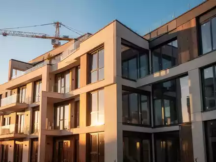 Ein modernes Mehrfamilienhaus mit klaren Linien und großen Fenstern, das im Sonnenlicht erstrahlt. Im Hintergrund ist ein Baukran zu sehen. - Foto: über boerse-global.de