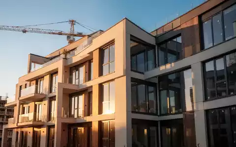 Ein modernes Mehrfamilienhaus mit klaren Linien und großen Fenstern, das im Sonnenlicht erstrahlt. Im Hintergrund ist ein Baukran zu sehen. - Foto: über boerse-global.de