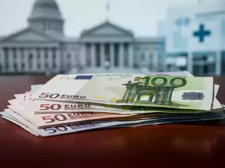 Nahaufnahme von gestapelten Euro-Banknoten auf einem dunklen Holztisch, mit verschwommenem Hintergrund eines Regierungsgebäudes. - Foto: über boerse-global.de