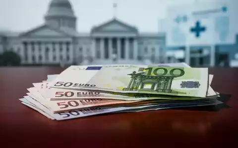 Nahaufnahme von gestapelten Euro-Banknoten auf einem dunklen Holztisch, mit verschwommenem Hintergrund eines Regierungsgebäudes. - Foto: über boerse-global.de