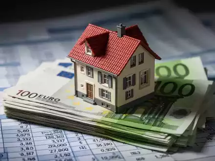 Ein detailliertes Miniaturhausmodell steht auf einem Stapel deutscher Euro-Banknoten, im Hintergrund verschwommene Finanzdokumente. - Foto: über boerse-global.de