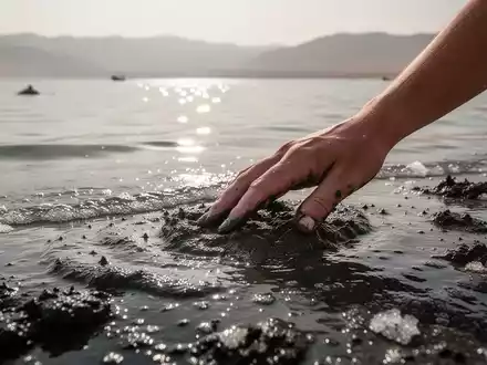 Eine Hand berührt den mineralreichen schwarzen Schlamm des Toten Meeres, mit dem Wasser und den Bergen im Hintergrund. - Foto: über boerse-global.de