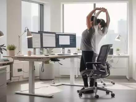 Ein ergonomischer Arbeitsplatz mit höhenverstellbarem Schreibtisch, ergonomischem Stuhl und einer Person, die sich streckt. - Foto: über boerse-global.de