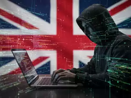 Eine Person im Kapuzenpulli tippt auf einem Laptop, umgeben von leuchtendem Code und Datenströmen, vor einer verschwommenen britischen Flagge. - Foto: über boerse-global.de