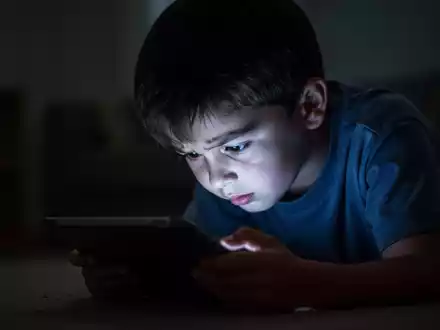 Ein Kind sitzt im Dunkeln, gebeugt über ein leuchtendes Tablet, dessen Licht sein Gesicht beleuchtet und die schlechte Haltung hervorhebt. - Foto: über boerse-global.de