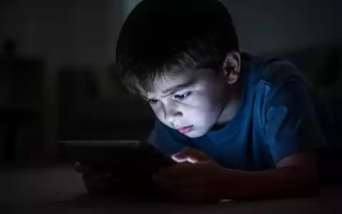 Ein Kind sitzt im Dunkeln, gebeugt über ein leuchtendes Tablet, dessen Licht sein Gesicht beleuchtet und die schlechte Haltung hervorhebt. - Foto: über boerse-global.de