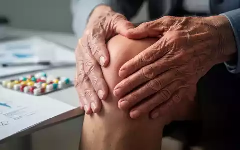 Ältere Hände berühren ein Knie, mit verschwommenen Nahrungsergänzungsmitteln im Hintergrund, was Gelenkschmerzen und Behandlungen symbolisiert. - Foto: über boerse-global.de