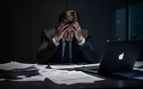 Eine Person sitzt mit den Händen am Kopf an einem Schreibtisch, umgeben von Dokumenten und einem Laptop, und wirkt überfordert. - Foto: über boerse-global.de