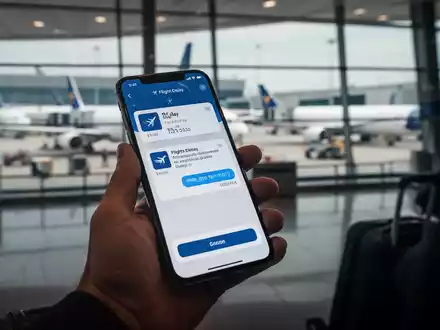 Eine Hand hält ein Smartphone mit einer Flugverspätungsanzeige, im Hintergrund unscharf ein Flughafen. Symbolisiert Reiseverzögerungen und Rechtsansprüche. - Foto: über boerse-global.de