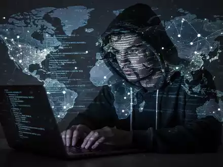 Schattenhafte Figur in Kapuzenpulli tippt auf Laptop, mit Code und Weltkarte als Projektionen, symbolisiert Cyber- und Georisiken. - Foto: über boerse-global.de
