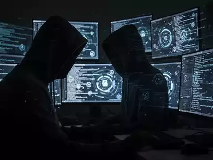 Eine dunkle, schattenhafte Figur sitzt vor mehreren leuchtenden Computermonitoren, die digitalen Code anzeigen. - Foto: über boerse-global.de