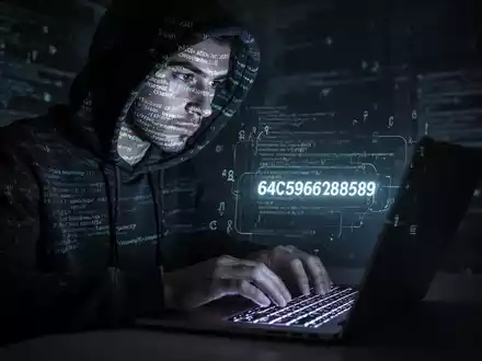 Eine Person im Kapuzenpulli tippt auf einer beleuchteten Tastatur, während Code und digitale Symbole auf ihr Gesicht projiziert werden. - Foto: über boerse-global.de