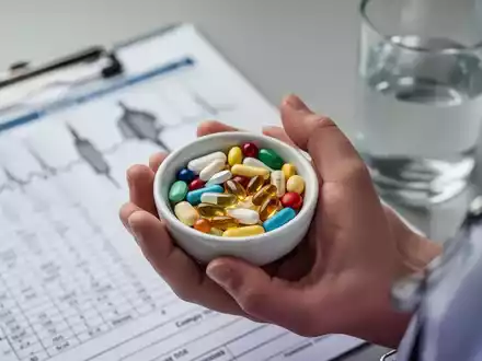 Nahaufnahme einer Hand, die eine Schale mit verschiedenen Vitaminpillen und Nahrungsergänzungsmitteln hält, im Hintergrund eine medizinische Tabelle. - Foto: über boerse-global.de