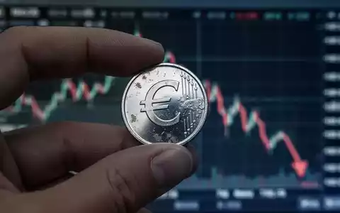 Eine Hand hält eine leicht angelaufene Silbermünze vor einem unscharfen, fallenden Börsenchart. Die Münze reflektiert ein Euro-Symbol. - Foto: über boerse-global.de