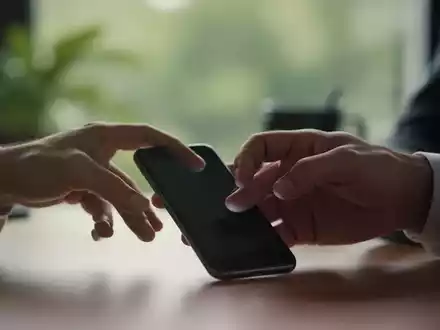 Eine Hand greift nach einem Smartphone, zögert aber, mit einem verschwommenen Hintergrund, der Ruhe oder einen Arbeitsplatz andeutet. - Foto: über boerse-global.de
