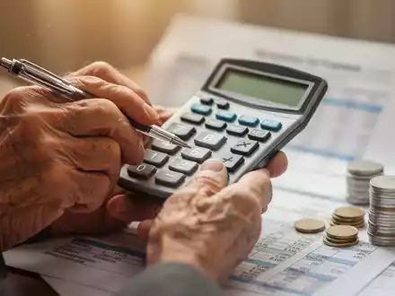 Nahaufnahme der Hände einer älteren Person, die einen Taschenrechner und einen Stift halten, mit verschwommenen Dokumenten und Münzen. - Foto: über boerse-global.de