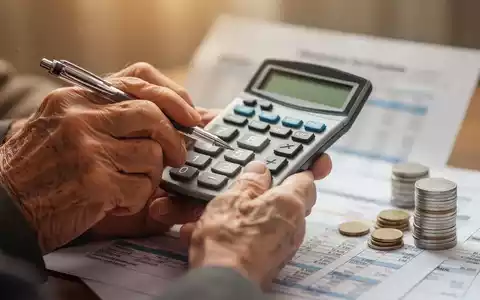 Nahaufnahme der Hände einer älteren Person, die einen Taschenrechner und einen Stift halten, mit verschwommenen Dokumenten und Münzen. - Foto: über boerse-global.de