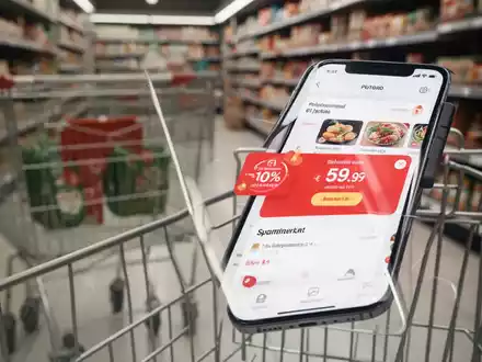Ein Smartphone mit einer Supermarkt-App, die Rabatte anzeigt, im Vordergrund; im Hintergrund verschwommen Supermarktregale. - Foto: über boerse-global.de