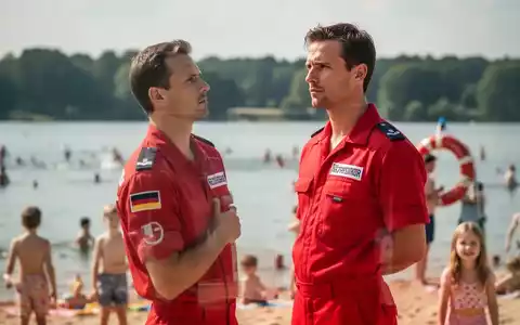 Ein Rettungsschwimmer in roter Uniform steht an einem belebten See oder Strand und blickt wachsam über das Wasser. - Foto: über boerse-global.de