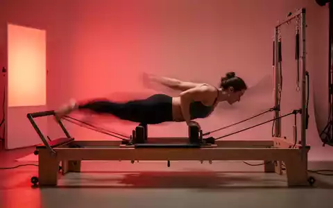 Eine Person auf einem Pilates Reformer, umgeben von warmem Rotlicht, das die Muskeln während des Trainings beleuchtet. - Foto: über boerse-global.de