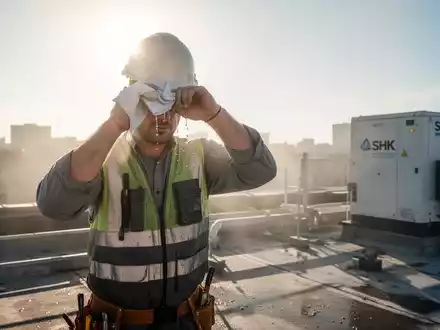 Ein Bauarbeiter wischt sich auf einem sonnenüberfluteten Dach den Schweiß von der Stirn, mit moderner SHK-Ausrüstung im Hintergrund. - Foto: über boerse-global.de