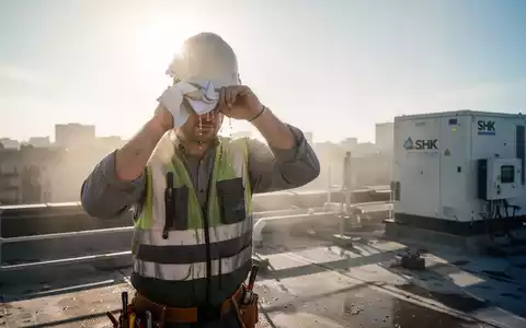 Ein Bauarbeiter wischt sich auf einem sonnenüberfluteten Dach den Schweiß von der Stirn, mit moderner SHK-Ausrüstung im Hintergrund. - Foto: über boerse-global.de