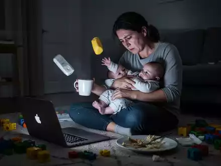 Eine erschöpfte Mutter arbeitet am Laptop, während sie ein Baby hält. Spielzeug und Geschirr liegen verstreut herum. - Foto: über boerse-global.de