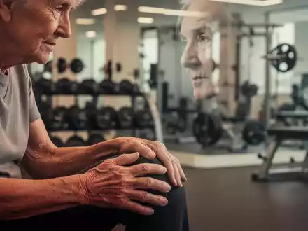 Ăltere Person berĂŒhrt ihr Knie, im Hintergrund ein moderner Physiotherapie-Fitnessraum mit GerĂ€ten. Fokus auf Gelenkschmerzen und Bewegung. - Foto: ĂŒber boerse-global.de
