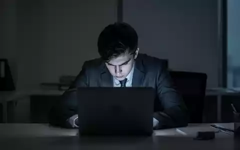 Eine junge Person sitzt gebeugt vor einem Laptop in einem dunklen Büro, symbolisierend stillen Stress und Überlastung. - Foto: über boerse-global.de