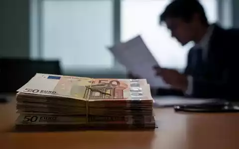 Gestapelte Euro-Banknoten auf einem Schreibtisch, unscharf, im Hintergrund eine Person, die Dokumente prüft. Symbolisiert finanzielle Belastung und politische Debatten im Gesundheitswesen. - Foto: über boerse-global.de