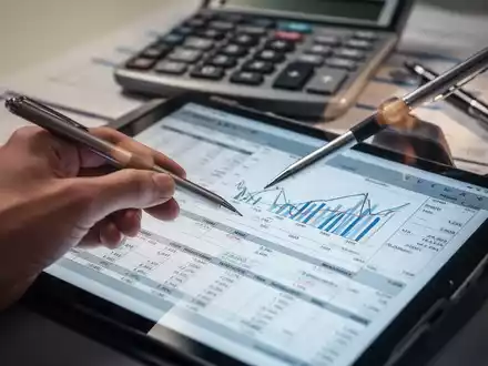 Hand hält Stift, zeigt auf Finanztabelle auf Tablet, mit Taschenrechner und Stift im Hintergrund. - Foto: über boerse-global.de