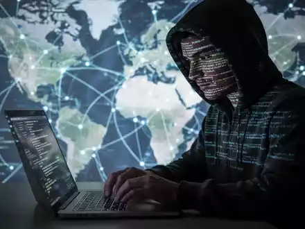Verschwommene Gestalt in Kapuzenjacke tippt auf Laptop, Code spiegelt sich im Gesicht, unscharfe Karte im Hintergrund. - Foto: über boerse-global.de
