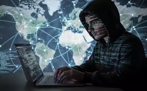 Verschwommene Gestalt in Kapuzenjacke tippt auf Laptop, Code spiegelt sich im Gesicht, unscharfe Karte im Hintergrund. - Foto: über boerse-global.de