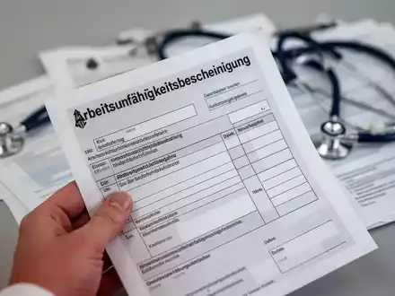 Eine Hand hält ein deutsches Formular für eine Arbeitsunfähigkeitsbescheinigung, mit verschwommenem Stethoskop im Hintergrund. - Foto: über boerse-global.de