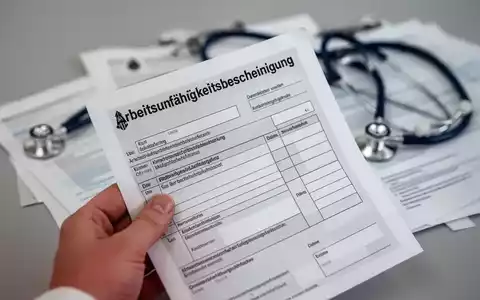 Eine Hand hält ein deutsches Formular für eine Arbeitsunfähigkeitsbescheinigung, mit verschwommenem Stethoskop im Hintergrund. - Foto: über boerse-global.de