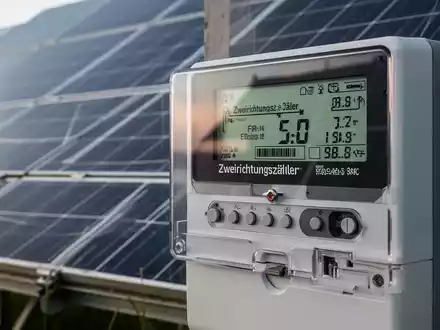 Ein moderner Zweirichtungszähler mit digitaler Anzeige, der Energieflussdaten darstellt, mit einer Solaranlage im Hintergrund. - Foto: über boerse-global.de