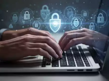 Nahaufnahme von Händen, die auf einer Laptop-Tastatur tippen, mit unscharfen digitalen Sicherheitssymbolen im Hintergrund. - Foto: über boerse-global.de