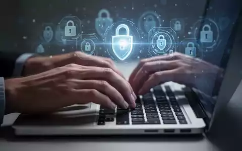 Nahaufnahme von Händen, die auf einer Laptop-Tastatur tippen, mit unscharfen digitalen Sicherheitssymbolen im Hintergrund. - Foto: über boerse-global.de