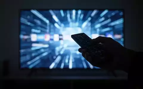 Eine Hand hält eine Fernbedienung, die auf einen unscharfen, leuchtenden Fernsehbildschirm mit digitalen Mustern zeigt. - Foto: über boerse-global.de