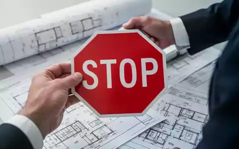 Eine Hand hält ein rotes Stoppschild vor verschwommenen Bauplänen und Dokumenten, symbolisierend das Ende der Gebäudeförderung. - Foto: über boerse-global.de