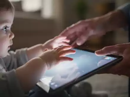 Eine Nahaufnahme von Kinderhänden, die nach einem leuchtenden Tablet greifen, während eine Erwachsenenhand versucht, sie wegzulenken. - Foto: über boerse-global.de