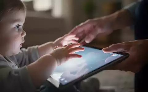 Eine Nahaufnahme von Kinderhänden, die nach einem leuchtenden Tablet greifen, während eine Erwachsenenhand versucht, sie wegzulenken. - Foto: über boerse-global.de