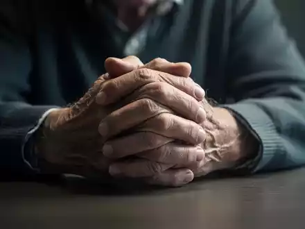 Nahaufnahme der Hände einer älteren Person, die besorgt auf einem Tisch gefaltet sind, mit weichem, unsicherem Licht. - Foto: über boerse-global.de