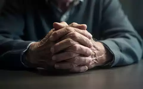 Nahaufnahme der Hände einer älteren Person, die besorgt auf einem Tisch gefaltet sind, mit weichem, unsicherem Licht. - Foto: über boerse-global.de